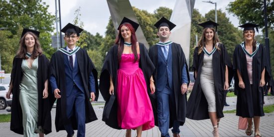 Six graduates walking in a row, girls in a pink dress in the middle